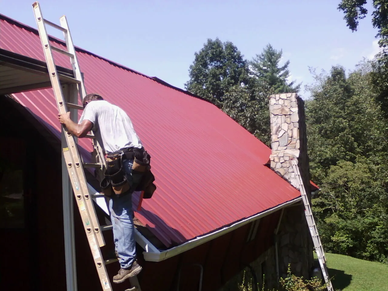 Roofer on a ladder beside a red metal roof and stone chimney — Soffit & Fascia Repair by Roofing Brothers in Helmville
